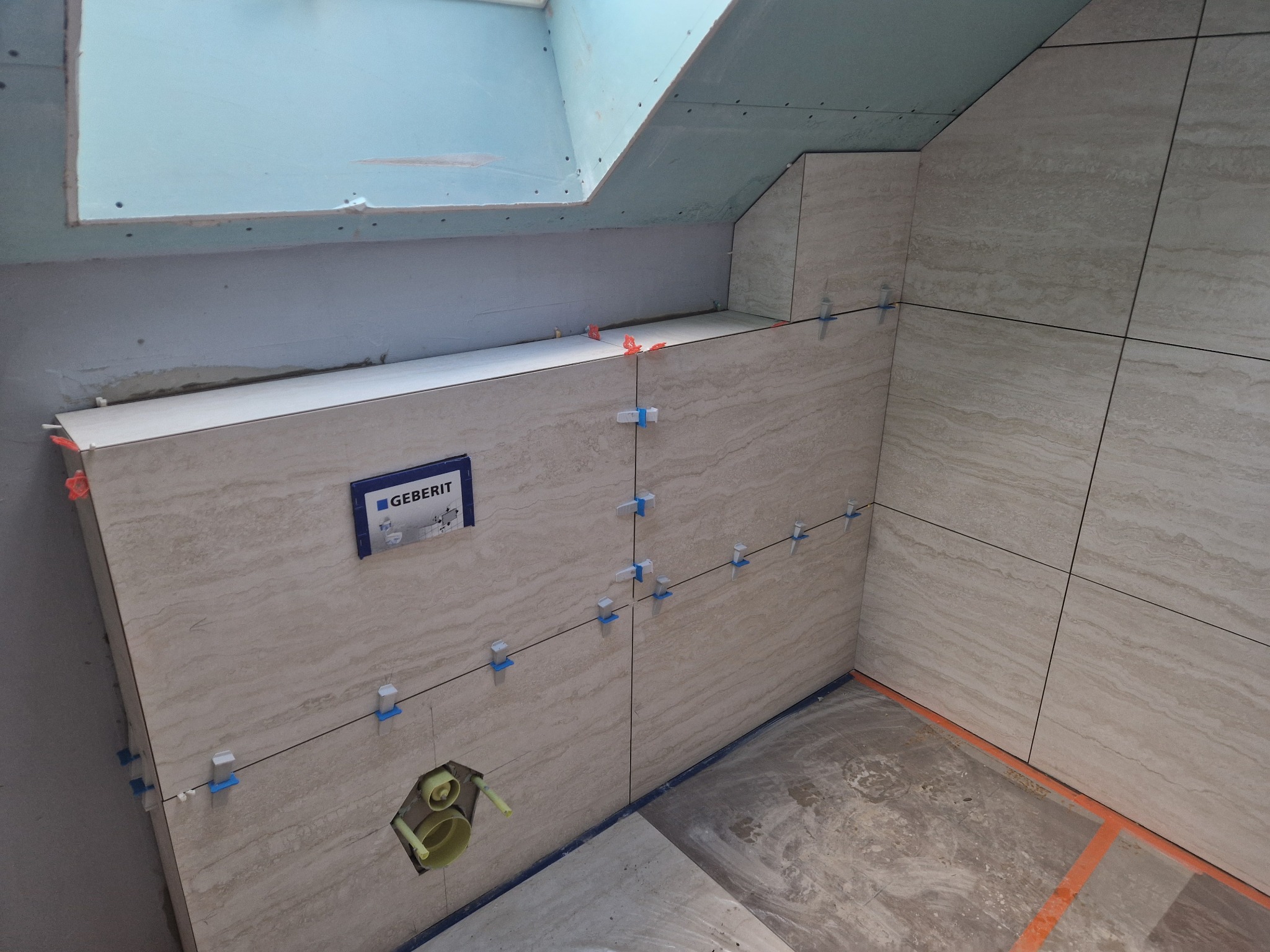 Ściana łazienki w trakcie wykańczania, z beżowymi płytkami imitującymi kamień, krzyżyki dystansowe, widoczna instalacja podtynkowa Geberit i sufit z płyt karton-gips.