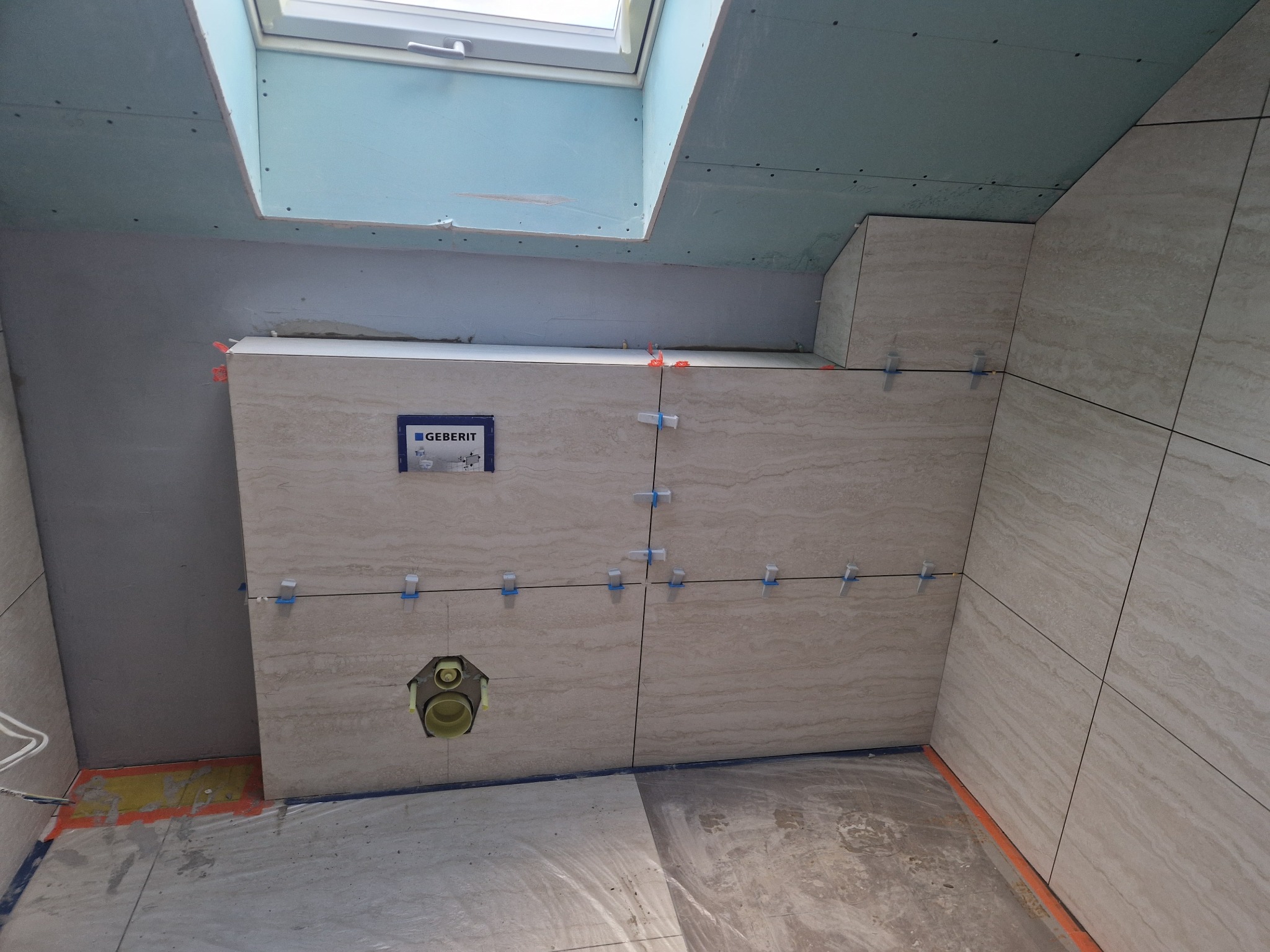 Wnętrze łazienki w trakcie prac glazurniczych: beżowe płytki imitujące trawertyn, krzyżyki dystansowe, widoczny stelaż podtynkowy Geberit i okno dachowe.