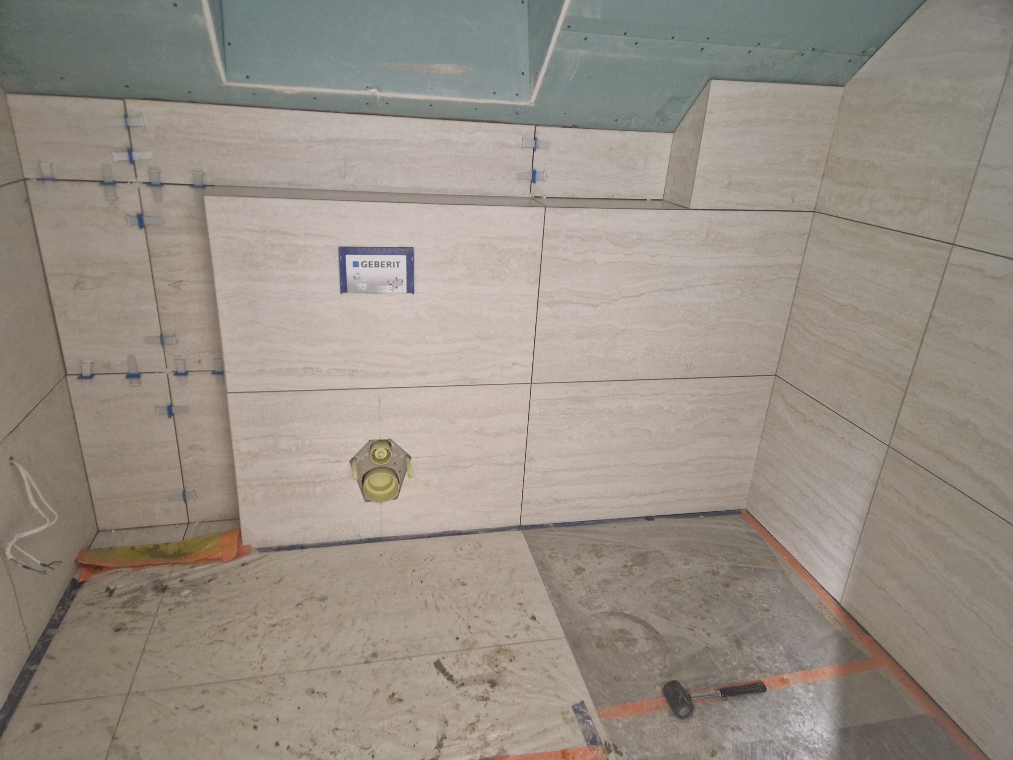 Wnętrze łazienki w trakcie prac: beżowe płytki imitujące kamień na ścianach, widoczne elementy instalacji Geberit i narzędzia glazurnicze. Podłoga częściowo wyłożona płytkami.