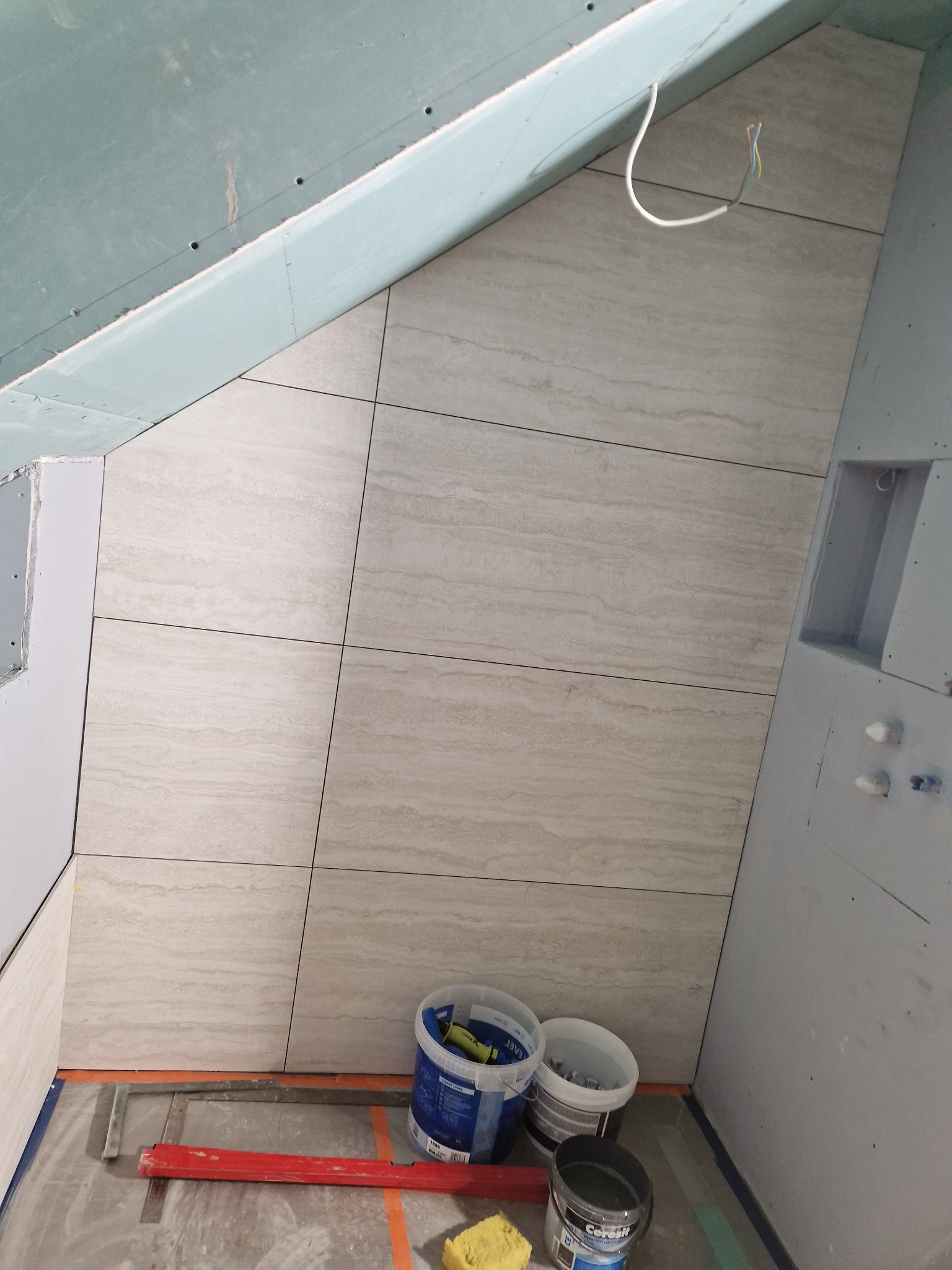 Ściana w skosie wyłożona beżowymi płytkami imitującymi trawertyn, widoczne narzędzia i materiały budowlane, sufit z płyt kartonowo-gipsowych w trakcie prac wykończeniowych.