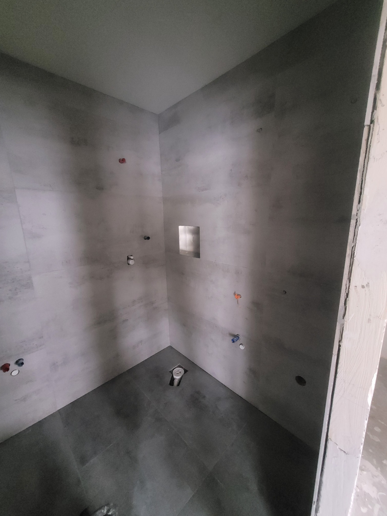 Wnętrze łazienki w trakcie remontu: szare płytki ścienne i podłogowe, widoczne przyłącza wodne i odpływ podłogowy, surowy ościeżnica drzwiowa.