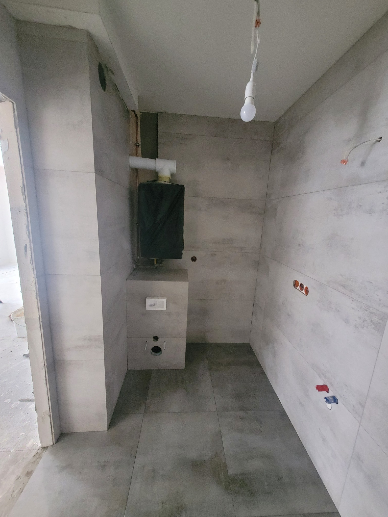 Wnętrze łazienki w trakcie remontu: szare płytki na ścianach i podłodze, zamaskowany piec CO, wystające kable elektryczne i rury wodne.
