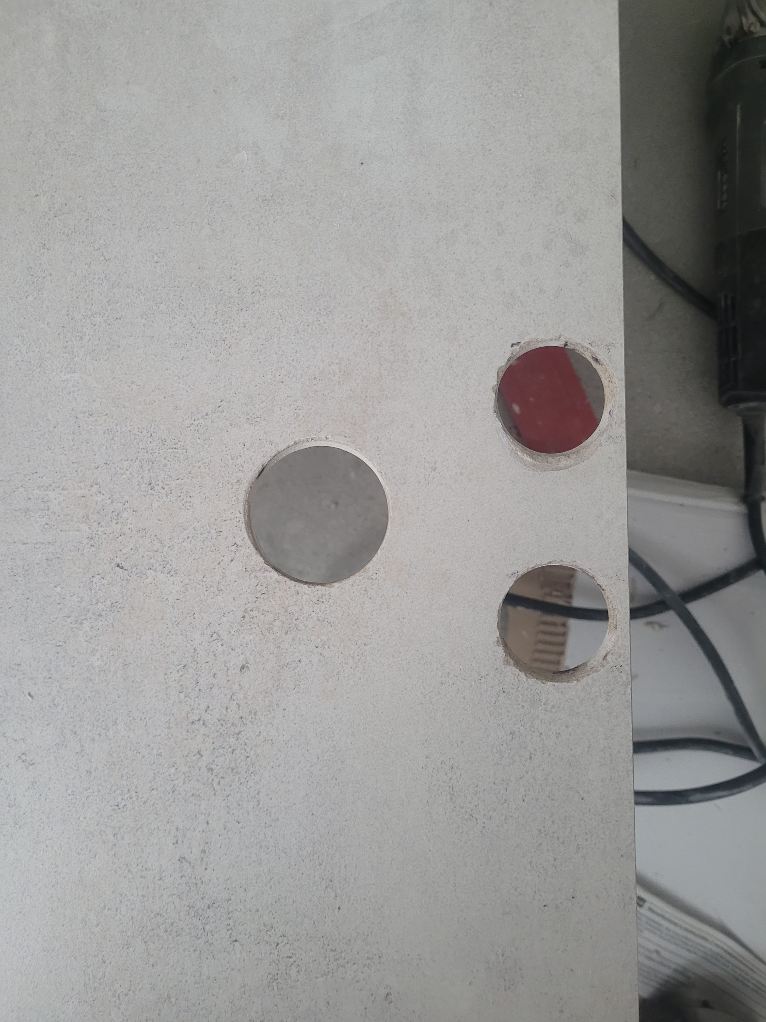 Płyta z jasnoszarej terakoty z trzema wywierconymi otworami o różnej średnicy, widoczne narzędzie do wiercenia obok.