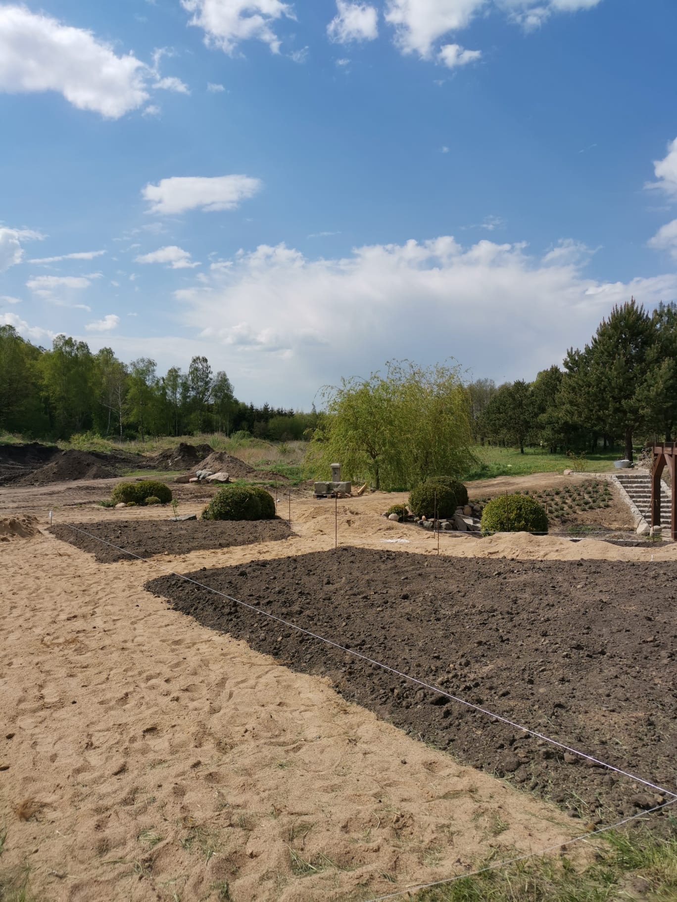 Przygotowanie terenu pod ogród: wytyczone grządki z ciemną ziemią na piaskowym podłożu, w tle drzewa i błękitne niebo z chmurami.