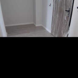 Piotr Drabik - Pomalowane na szaro wnętrze pokoju z nową, drewnianą ościeżnicą drzwiową zabezpieczoną folią ochronną i zamontowaną klamką.