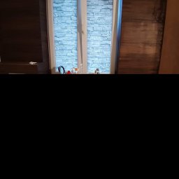 Piotr Drabik - Wnętrze pokoju z oknem, w którym zamontowano rolety imitujące kamienną ścianę, obok ściana wykończona panelami drewnopodobnymi.