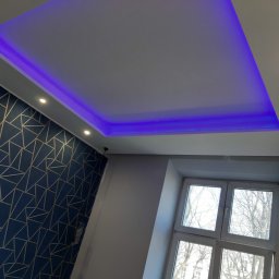 Piotr Drabik - Pokój po remoncie: Podwieszany sufit z oświetleniem LED w kolorze fioletowym, geometryczna tapeta w odcieniach granatu i złota, okno z widokiem na drzewa.