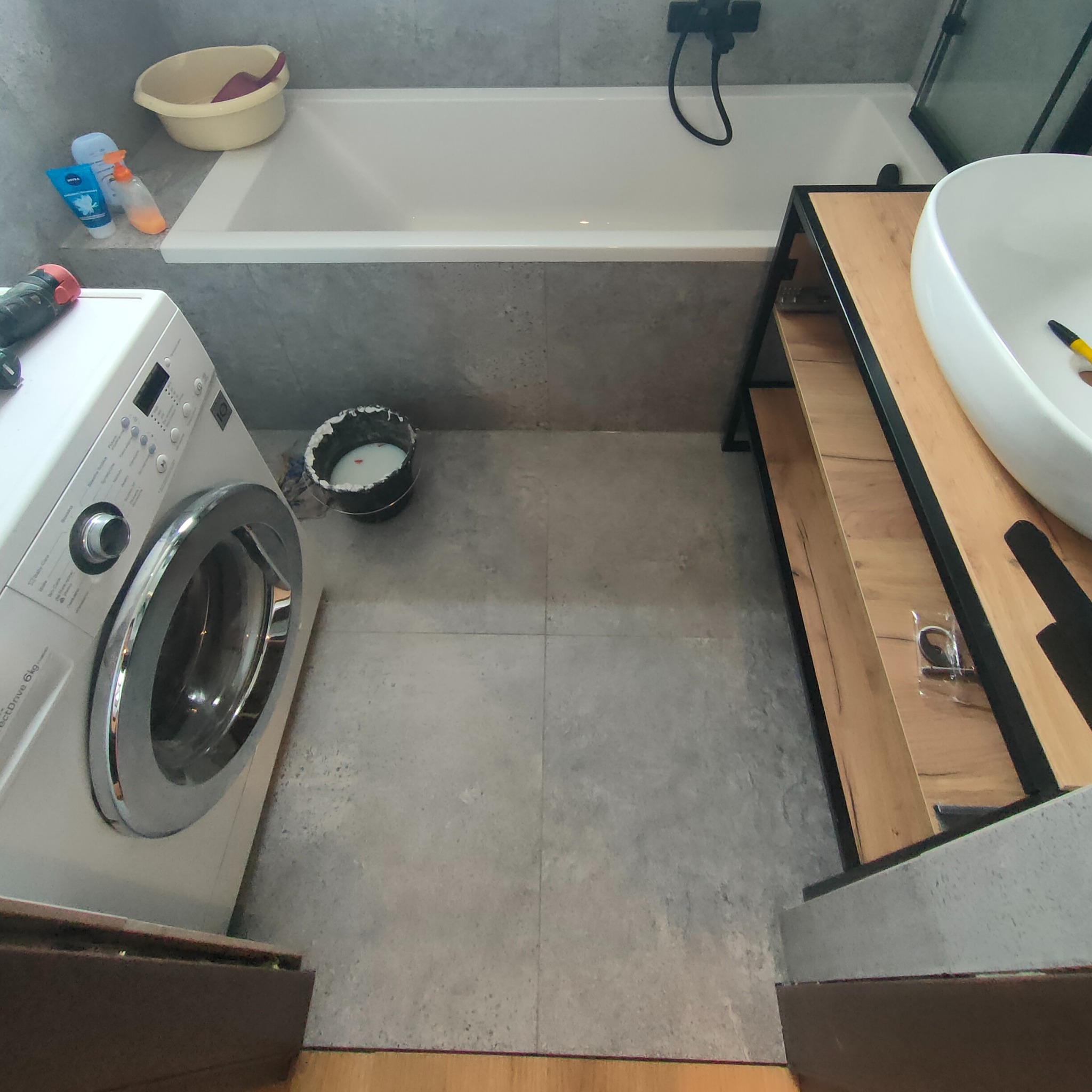 Łazienka w trakcie prac wykończeniowych; widoczna pralka, wanna, umywalka na drewnianym stelażu, szare płytki na podłodze i ścianach, wiadro z farbą oraz narzędzia.