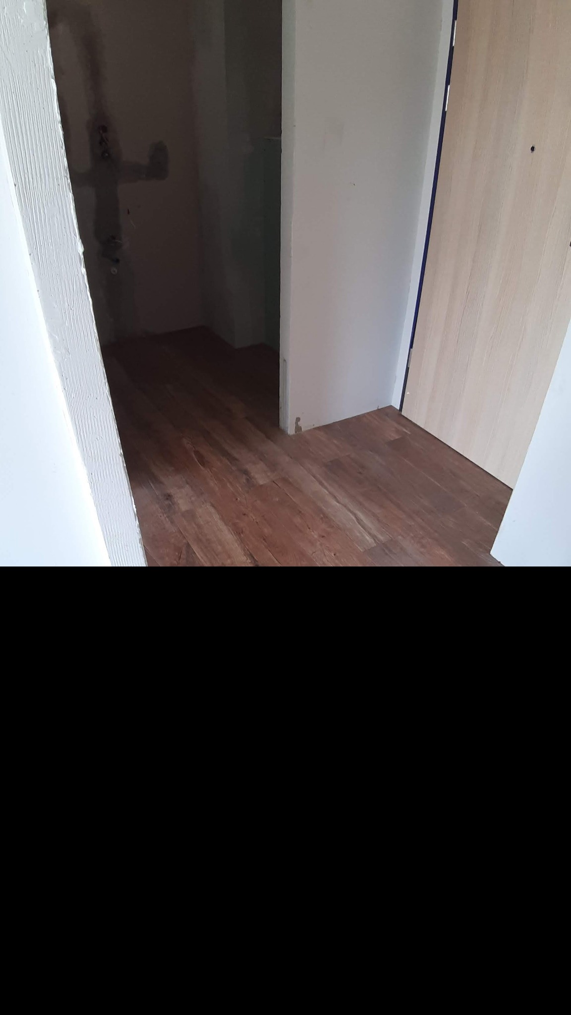 Ułożone panele podłogowe w odcieniach brązu, widoczne fragmenty ścian w kolorze białym i drzwi wewnętrzne z widoczną ościeżnicą, surowe ściany w głębi pomieszczenia z instalacją hydrauliczną.