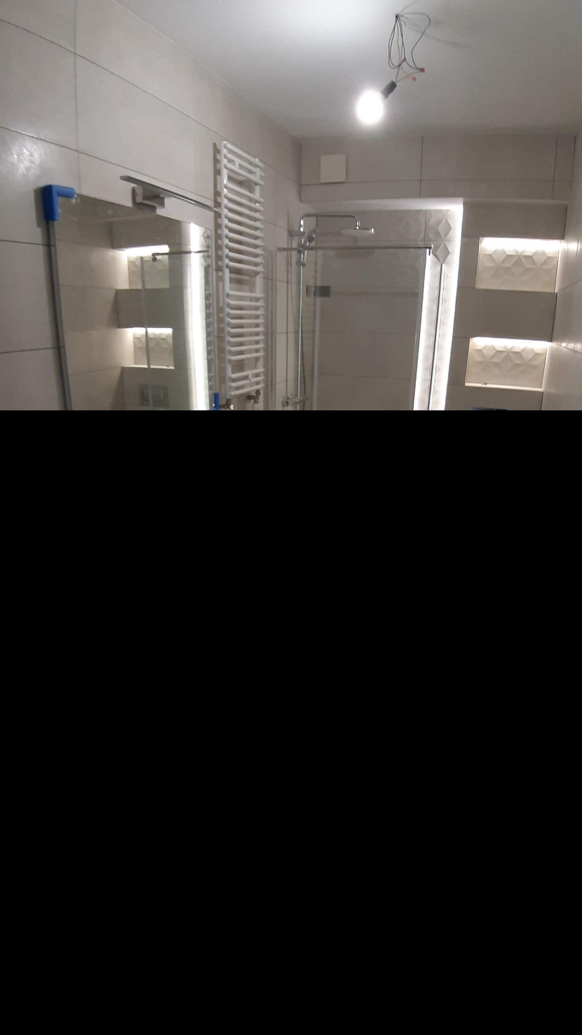 Nowoczesna łazienka z kabiną prysznicową, grzejnikiem drabinkowym, podświetlanymi wnękami i wiszącym lustrem. Widoczna żarówka na kablach zamiast lampy sufitowej.