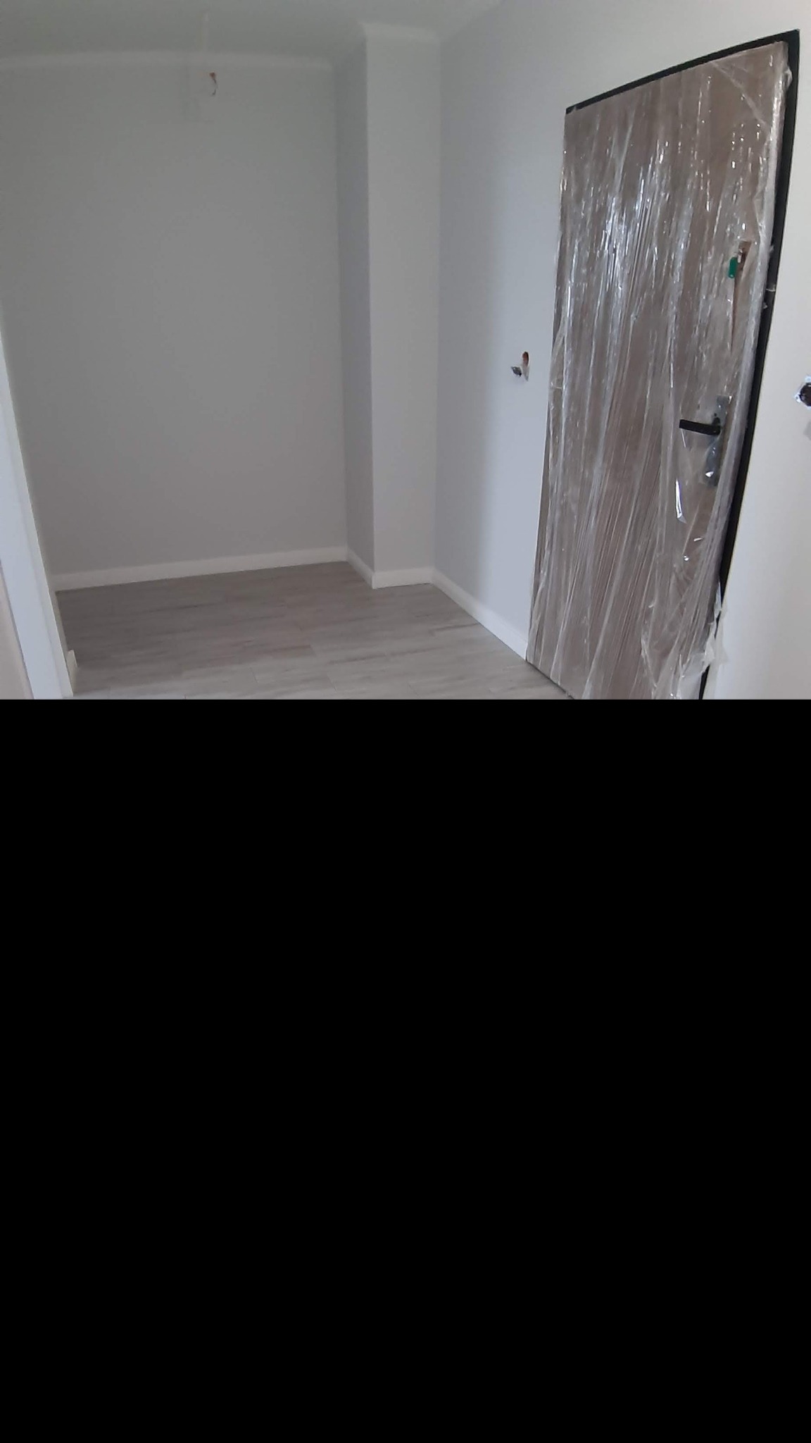 Pomalowane na szaro wnętrze pokoju z nową, drewnianą ościeżnicą drzwiową zabezpieczoną folią ochronną i zamontowaną klamką.