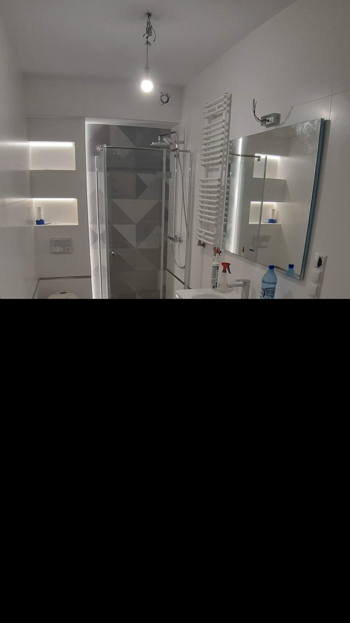 Nowoczesna łazienka z szarą kabiną prysznicową z geometrycznym wzorem, podświetlanymi wnękami w ścianie, grzejnikiem drabinkowym i dużym lustrem, widoczne niedokończone podłączenie oświetlenia...