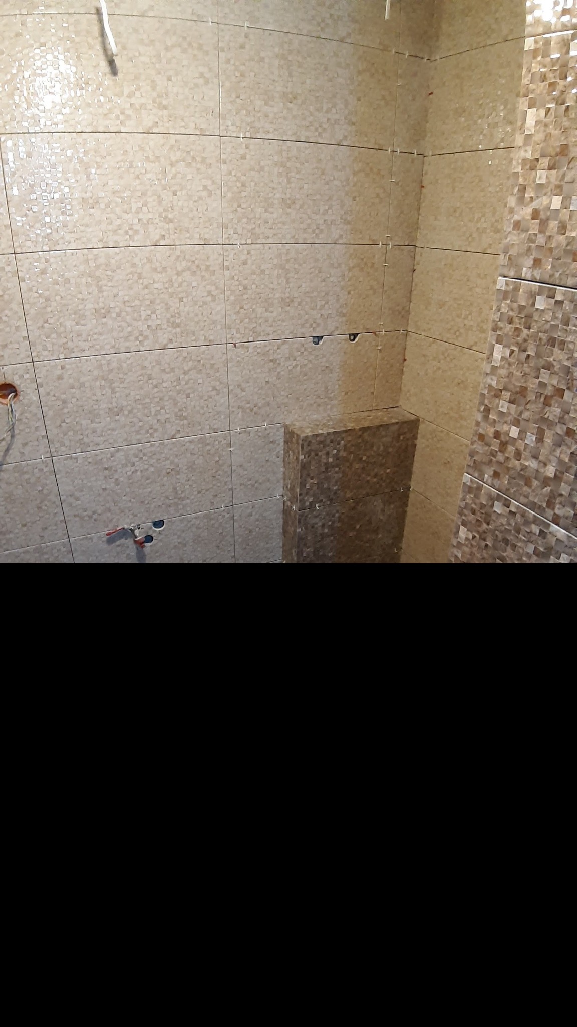 Ściana łazienki w trakcie układania beżowych płytek z drobną mozaiką, widoczne krzyżyki dystansowe i wystające rury instalacji wodnej.