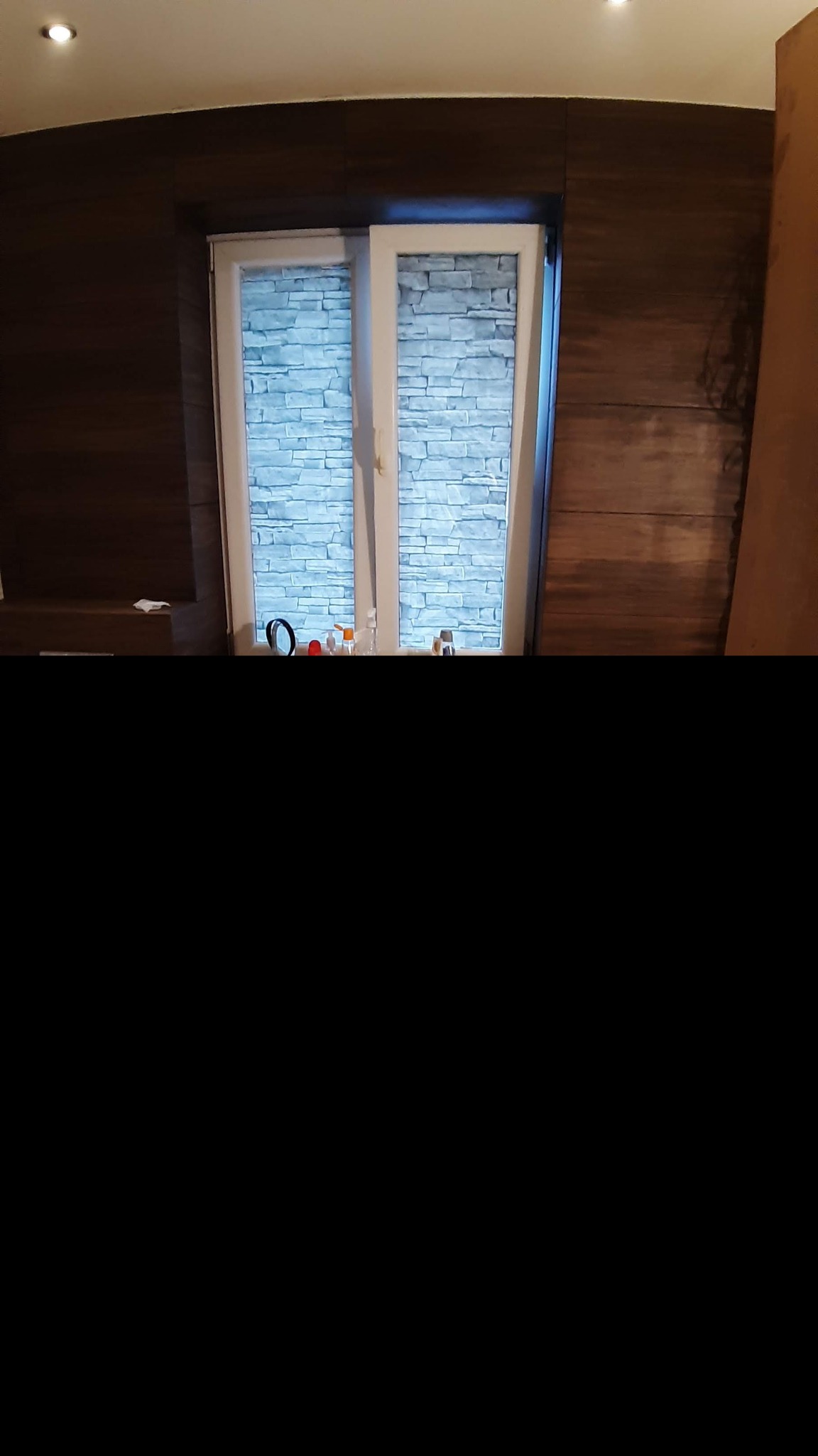 Wnętrze pokoju z oknem, w którym zamontowano rolety imitujące kamienną ścianę, obok ściana wykończona panelami drewnopodobnymi.