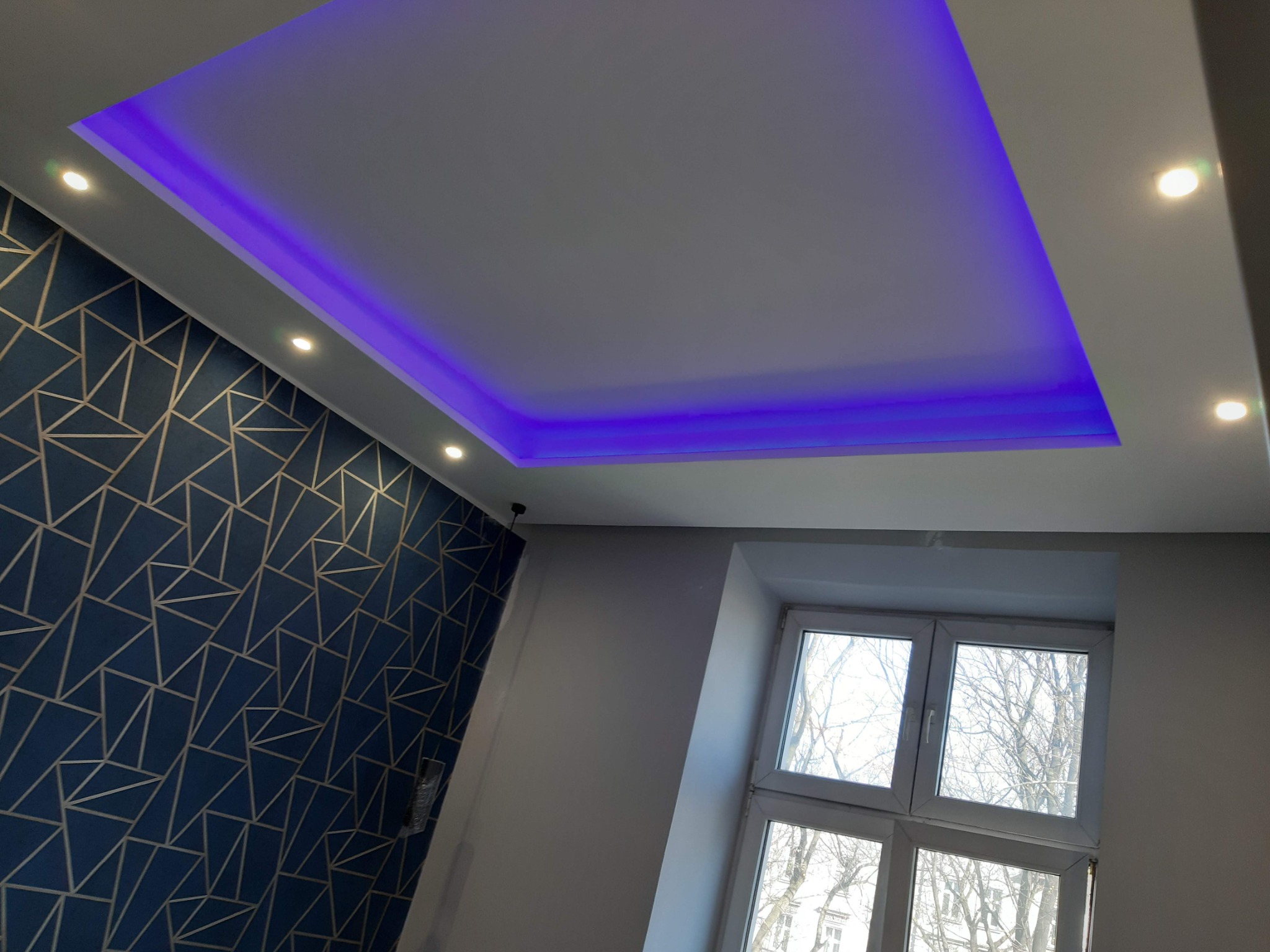 Pokój po remoncie: Podwieszany sufit z oświetleniem LED w kolorze fioletowym, geometryczna tapeta w odcieniach granatu i złota, okno z widokiem na drzewa.