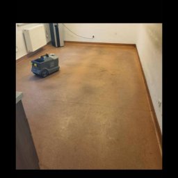 Mopik - Automat do mycia podłóg na brązowej, matowej posadzce w pustym pomieszczeniu biurowym, widoczny grzejnik i listwy przypodłogowe.