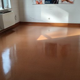 Mopik - Błyszcząca, brązowa podłoga po umyciu w pustym pokoju z białymi ścianami, kaloryferem i oknem po lewej stronie, oraz z kalendarzami na ścianie w tle.