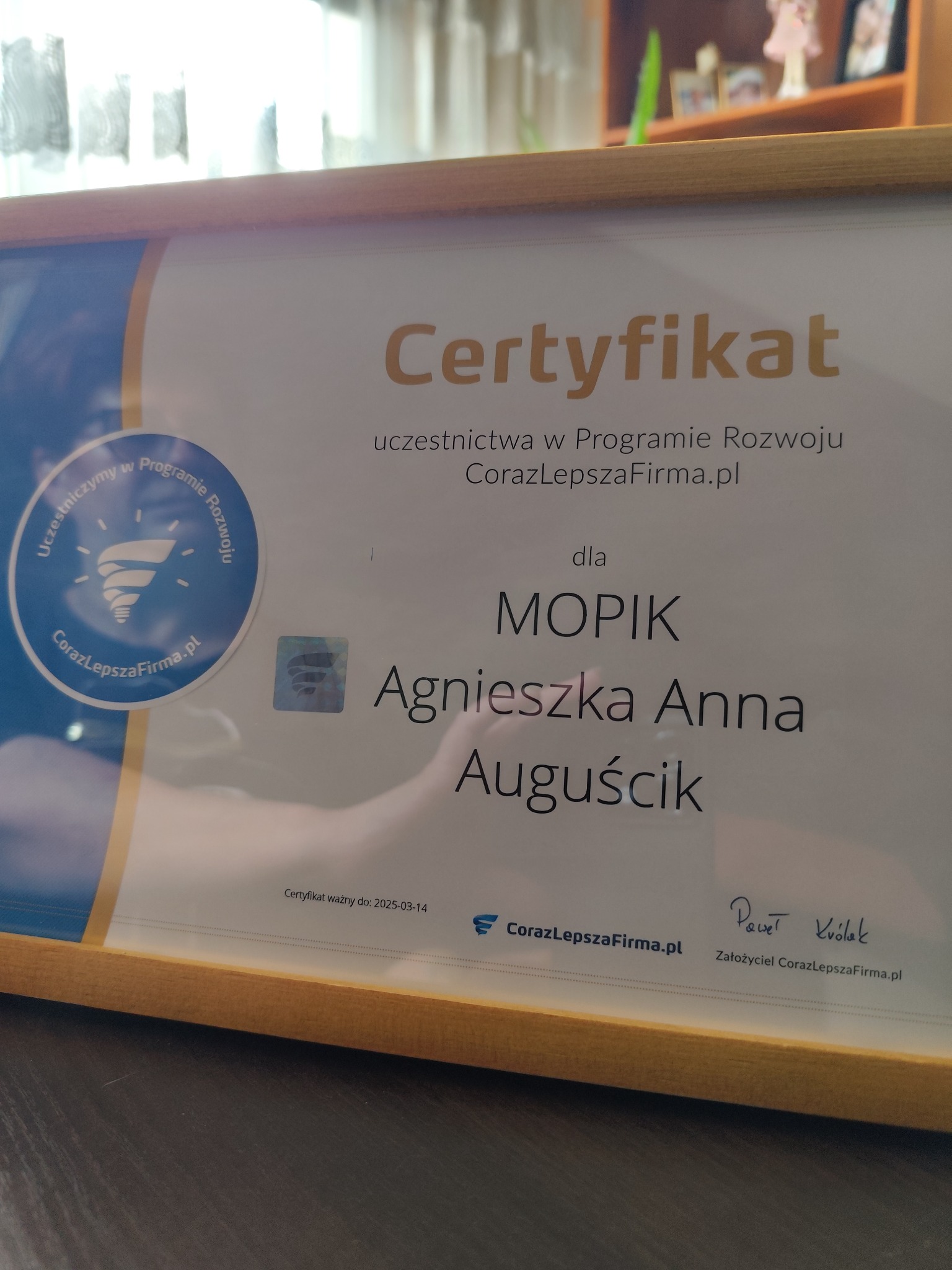 Oprawiony certyfikat uczestnictwa w programie rozwoju firmy CorazLepszaFirma.pl dla MOPIK Agnieszka Anna Auguścik, ważny do 2025-03-14, leżący na ciemnym blacie.