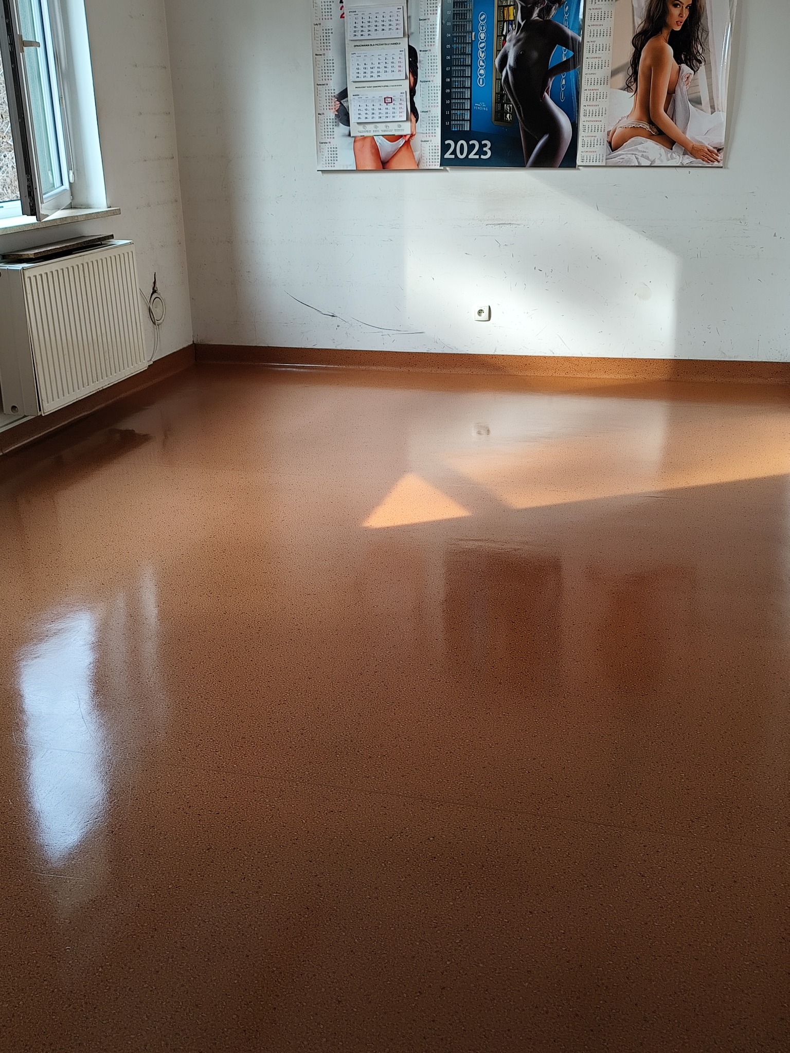Błyszcząca, brązowa podłoga po umyciu w pustym pokoju z białymi ścianami, kaloryferem i oknem po lewej stronie, oraz z kalendarzami na ścianie w tle.