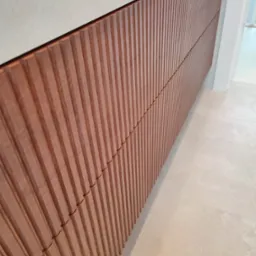 Nowoczesna zabudowa meblowa z ryflowanymi frontami w odcieniach brązu, widoczna z perspektywy podłogi, z jasnymi płytkami podłogowymi i fragmentem białej ściany w tle.