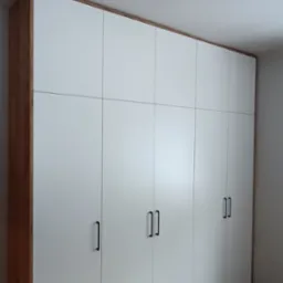 Duża szafa wnękowa z białymi frontami i drewnianym obramowaniem, z czarnymi, prostokątnymi uchwytami, na tle parkietu ułożonego w jodełkę.