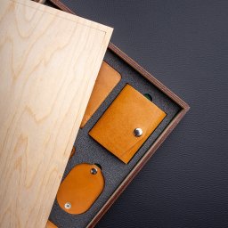 MokVok Sp. z O.O. - Zestaw galanterii skórzanej w drewnianym pudełku z jasnego drewna, zawierający portfelik zapinany na zatrzask, etui na karty i brelok, ułożony na czarnym tle.