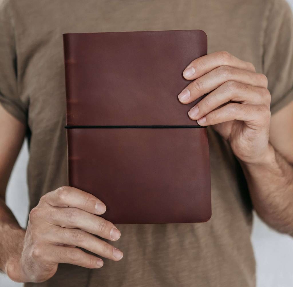 Osoba trzyma w dłoniach brązowy, skórzany notatnik z czarną gumką zamykającą.