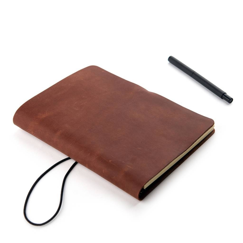 Brązowy skórzany notatnik z czarną elastyczną gumką i czarnym długopisem na białym tle.