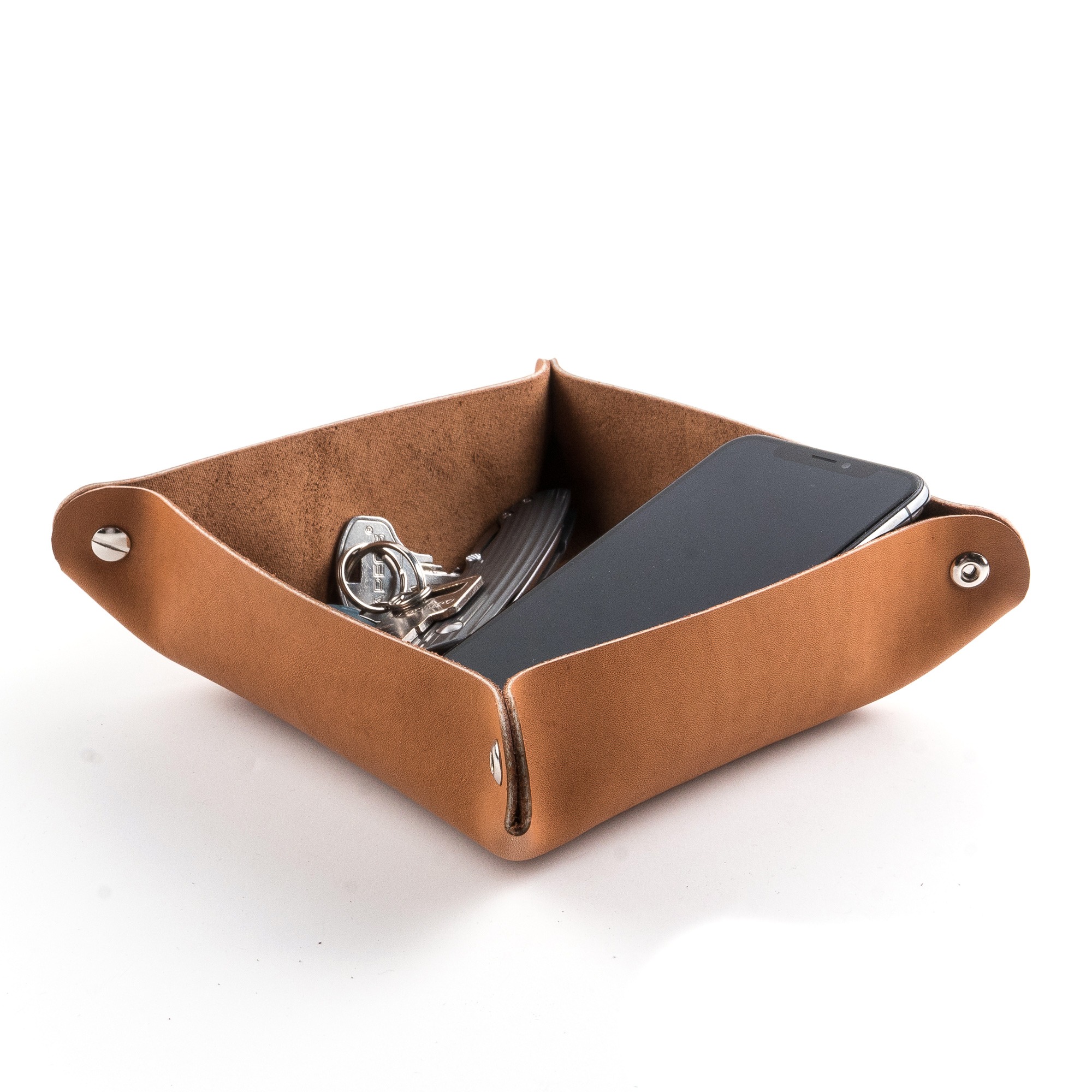 Skórzana tacka-organizer w kolorze karmelowym na białym tle, zawierająca telefon, klucze i scyzoryk. Tacka ma metalowe nity w rogach.
