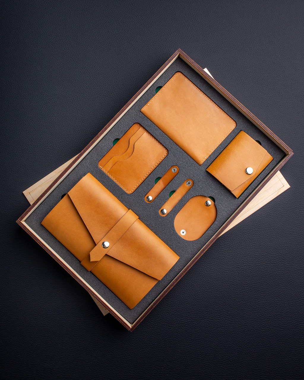 Zestaw galanterii skórzanej w odcieniach karmelowego brązu, zawierający portfel kopertowy, etui na karty, notes, mały portfelik i uchwyty na kable, umieszczony w drewnianym pudełku z szarą wkładką...