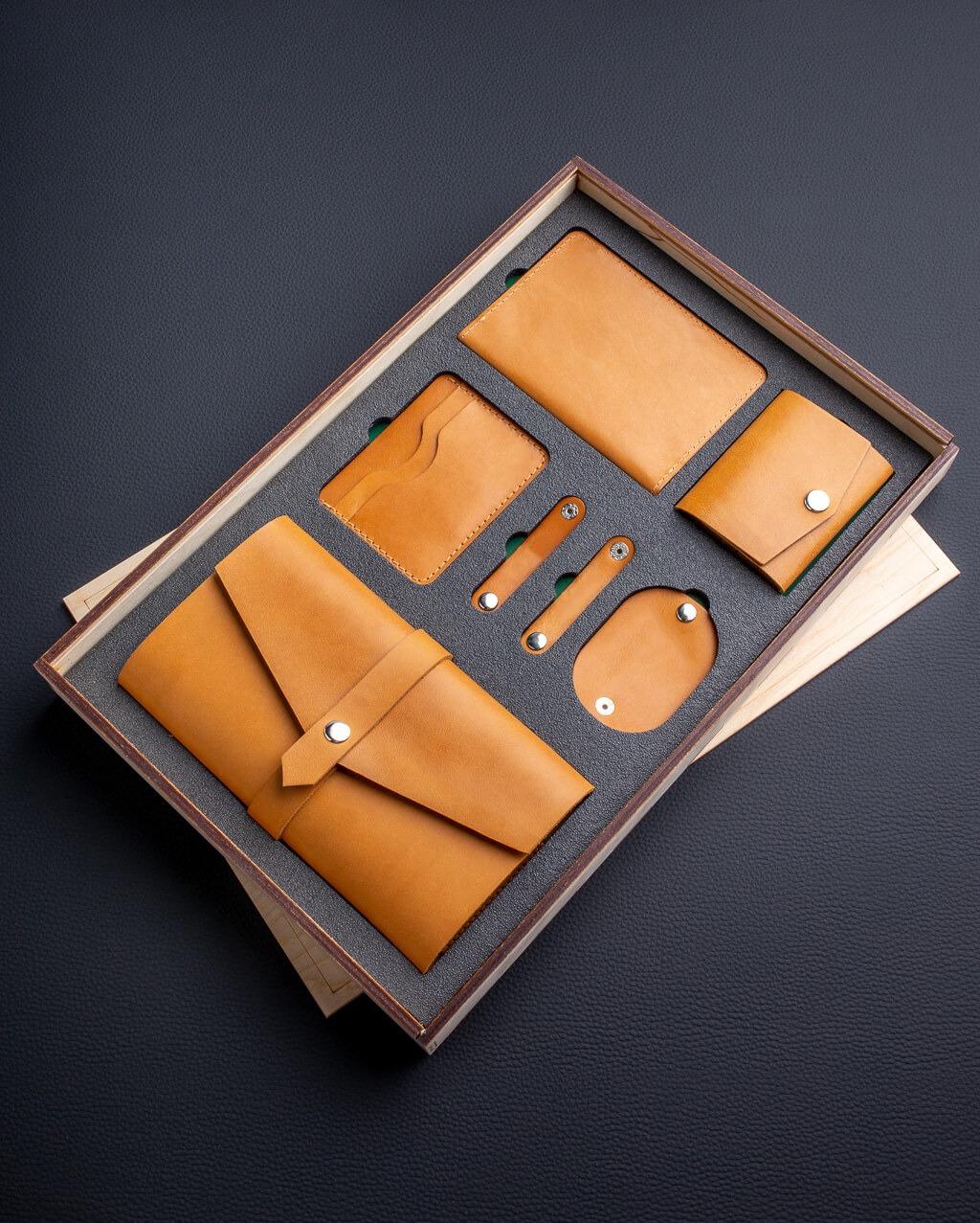 Zestaw galanterii skórzanej w drewnianym pudełku z szarą wkładką: portfel, etui na karty, okładka na dokumenty, mini portfel, uchwyty na kable. Wszystko w odcieniach brązu.
