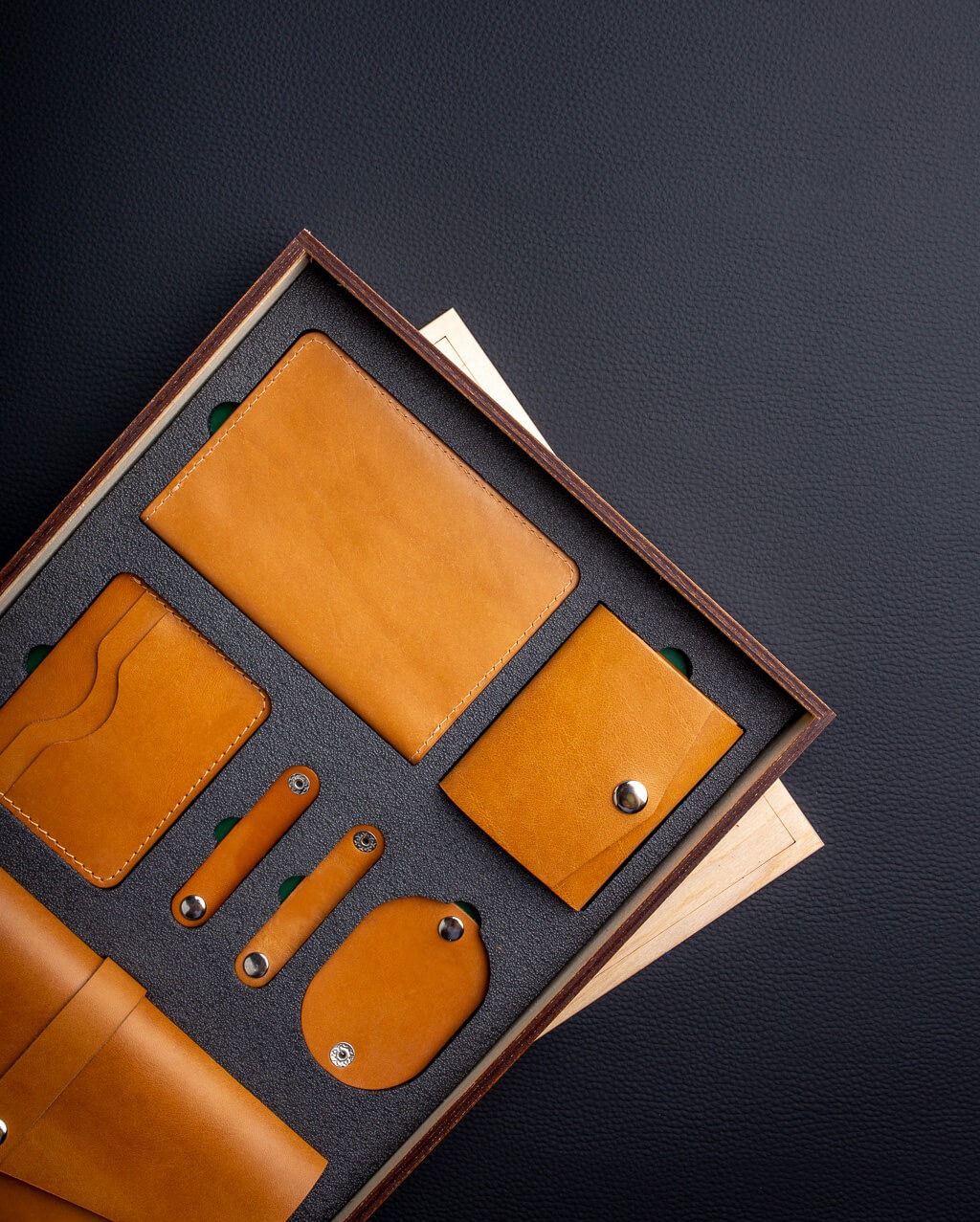 Zestaw galanterii skórzanej w drewnianym pudełku na czarnym tle, zawierający portfel, etui na karty, okładkę na dokumenty i uchwyty na kable, wszystko w odcieniach brązu i beżu.