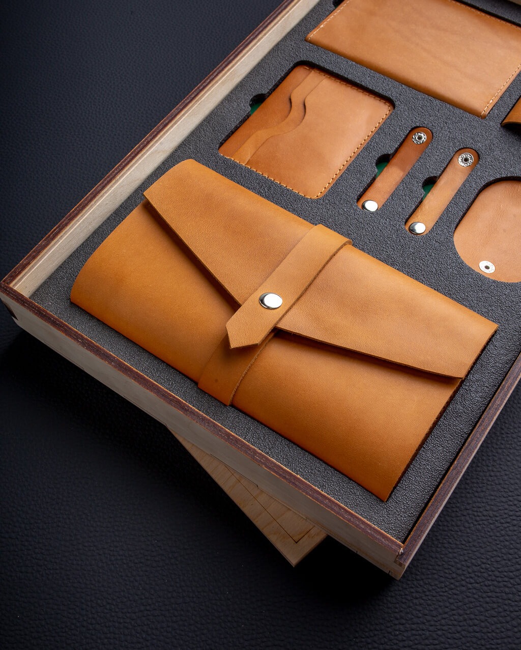 Zestaw galanterii skórzanej w drewnianym pudełku z szarym wypełnieniem: etui na dokumenty z zapięciem na zatrzask, portfel na karty, dwa uchwyty na kable z zatrzaskami i okrągły brelok, wszystko...