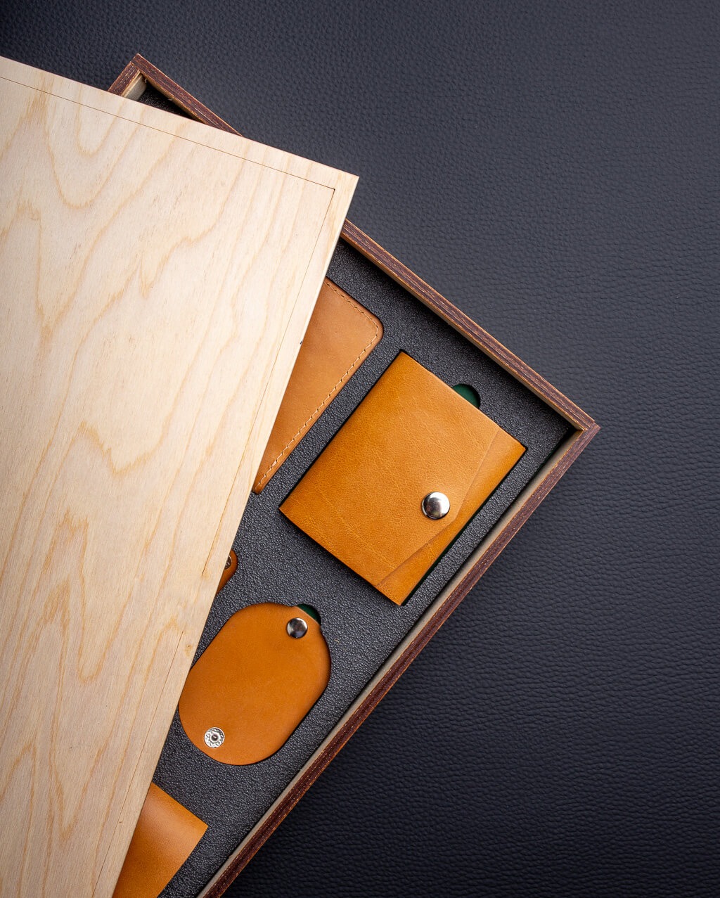 Zestaw galanterii skórzanej w drewnianym pudełku z jasnego drewna, zawierający portfelik zapinany na zatrzask, etui na karty i brelok, ułożony na czarnym tle.