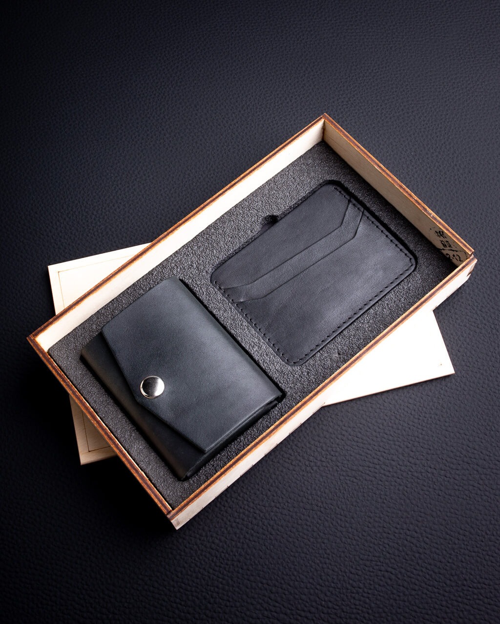 Zestaw skórzany: mini-portfel z zatrzaskiem i card holder w drewnianym pudełku, na czarnym, teksturowanym tle. Produkt dostępny w Piasecznie.