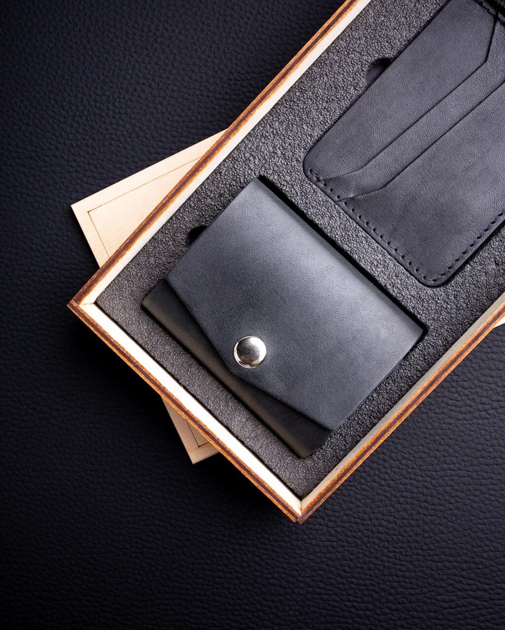 Zestaw galanterii skórzanej w drewnianym pudełku: minimalistyczny, dwupoziomowy card holder z zapięciem na zatrzask oraz etui na karty, wykończone stebnowaniem, na tle czarnej skóry.