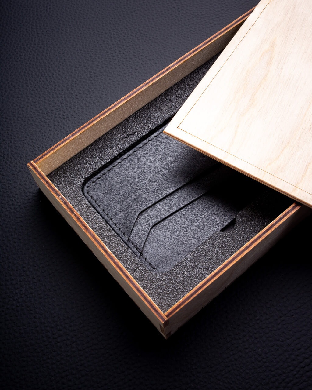Miniaturowy, czarny portfel skórzany typu card holder w drewnianym pudełku z wyściółką, widoczna część pokrywki, całość na ciemnym tle.