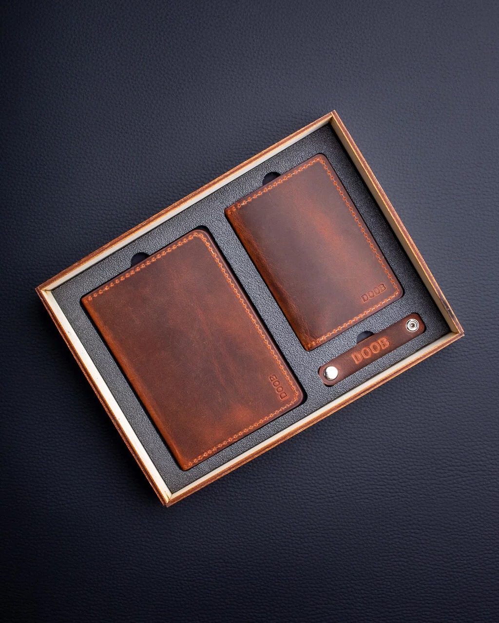 Zestaw prezentowy: skórzany organizer na dokumenty, mini-portfel i brelok z logo w drewnianym pudełku na czarnym tle.