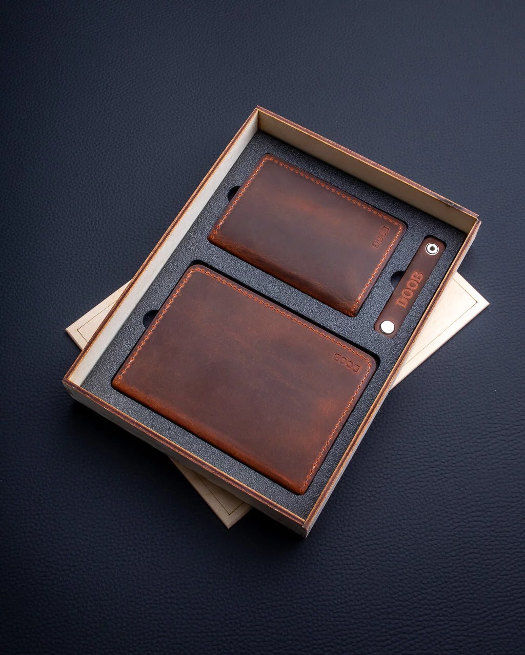 Zestaw galanterii skórzanej w drewnianym pudełku: portfel typu bifold, etui na karty i brelok z wygrawerowanym napisem, widoczne przeszycia i faktura skóry.