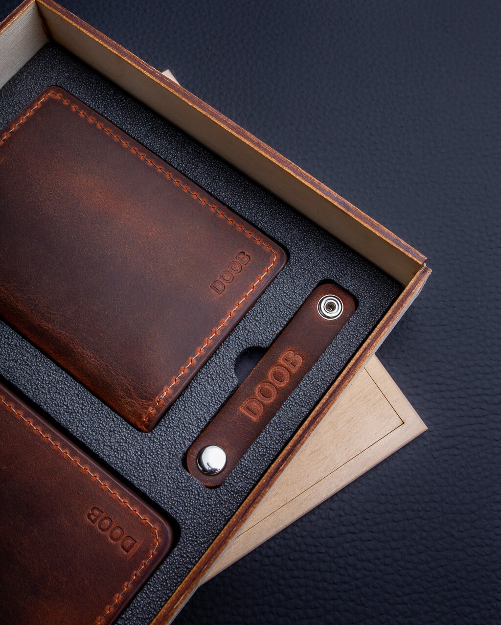 Zestaw prezentowy: brązowy skórzany portfel, etui na karty i brelok z metalowym nitem, umieszczone w pudełku z drewnianą wkładką na tle czarnej teksturowanej powierzchni.