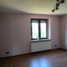 Łukasz Kuś - Pusty pokój z panelami podłogowymi w odcieniach brązu, ścianami w kolorze pudrowego różu, białym grzejnikiem pod oknem z roletami i metalową lampą sufitową.