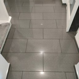 Łukasz Kuś - Szara, prostokątna podłoga z płytek ceramicznych w korytarzu, widok z góry, z fragmentem schodów i białej ościeżnicy drzwi.