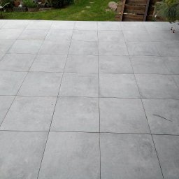 Łukasz Kuś - Układanie szarych, kwadratowych płytek na tarasie, widoczne pomarańczowe elementy poziomujące, w tle trawnik i drewniane ogrodzenie.