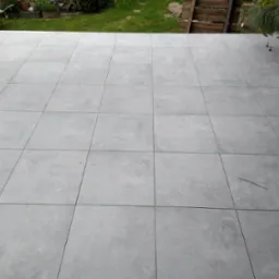 Układanie szarych, kwadratowych płytek na tarasie, widoczne pomarańczowe elementy poziomujące, w tle trawnik i drewniane ogrodzenie.