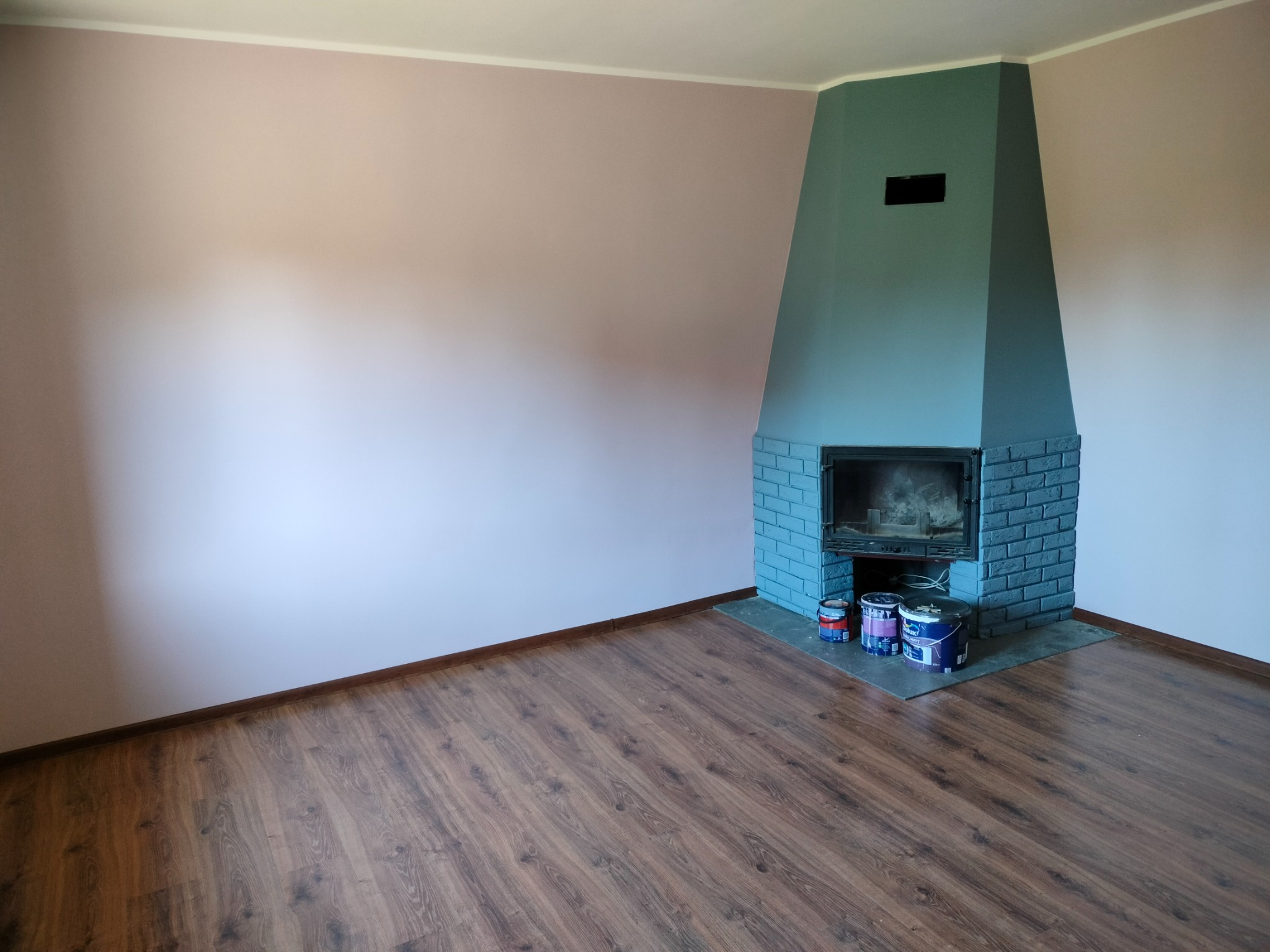 Pokój po remoncie: ściany pomalowane na różowo, kominek w odcieniach szarości, na podłodze panele imitujące drewno, widoczne puszki z farbą przy kominku.