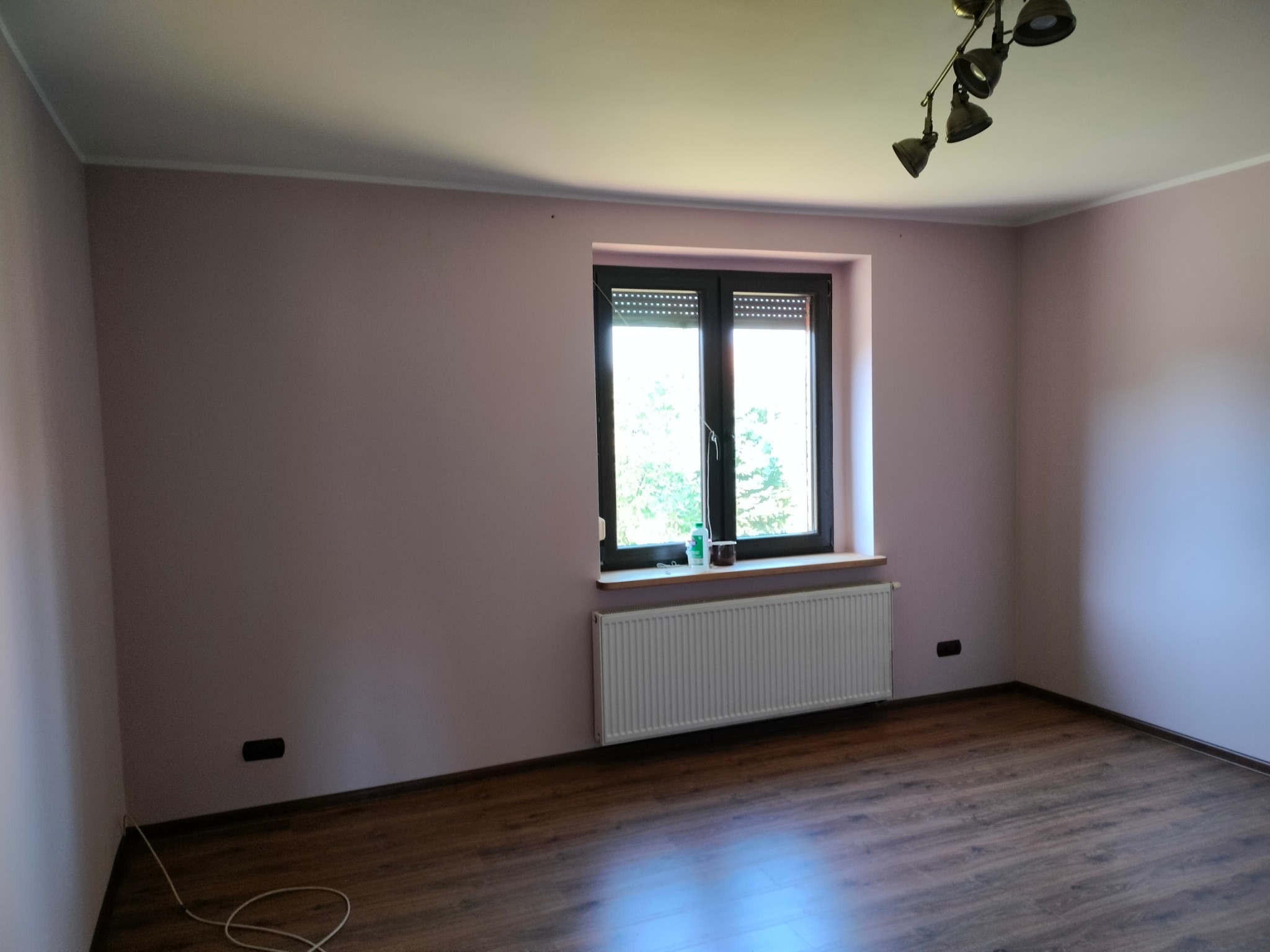Pusty pokój z panelami podłogowymi w odcieniach brązu, ścianami w kolorze pudrowego różu, białym grzejnikiem pod oknem z roletami i metalową lampą sufitową.