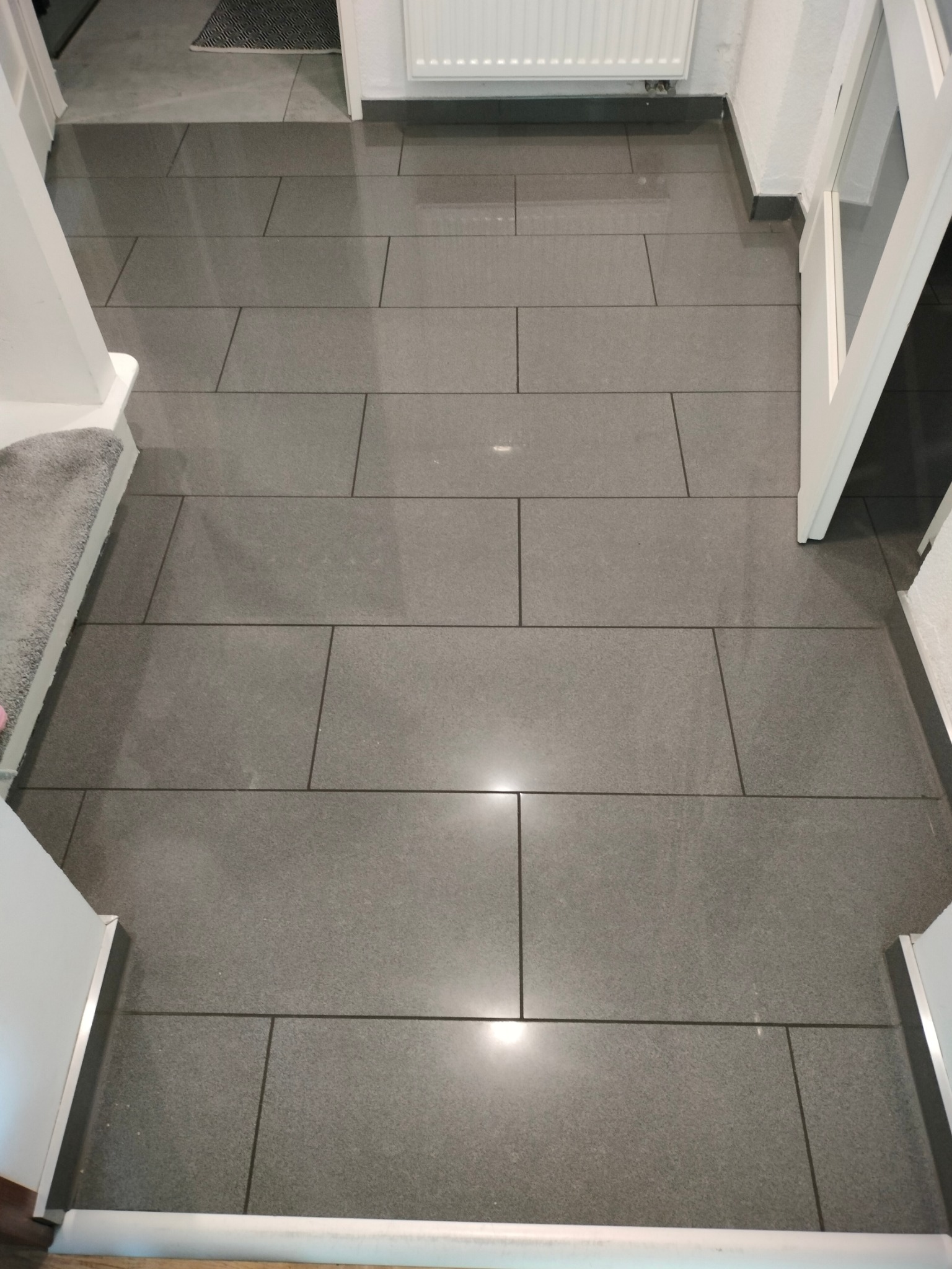 Szara, prostokątna podłoga z płytek ceramicznych w korytarzu, widok z góry, z fragmentem schodów i białej ościeżnicy drzwi.
