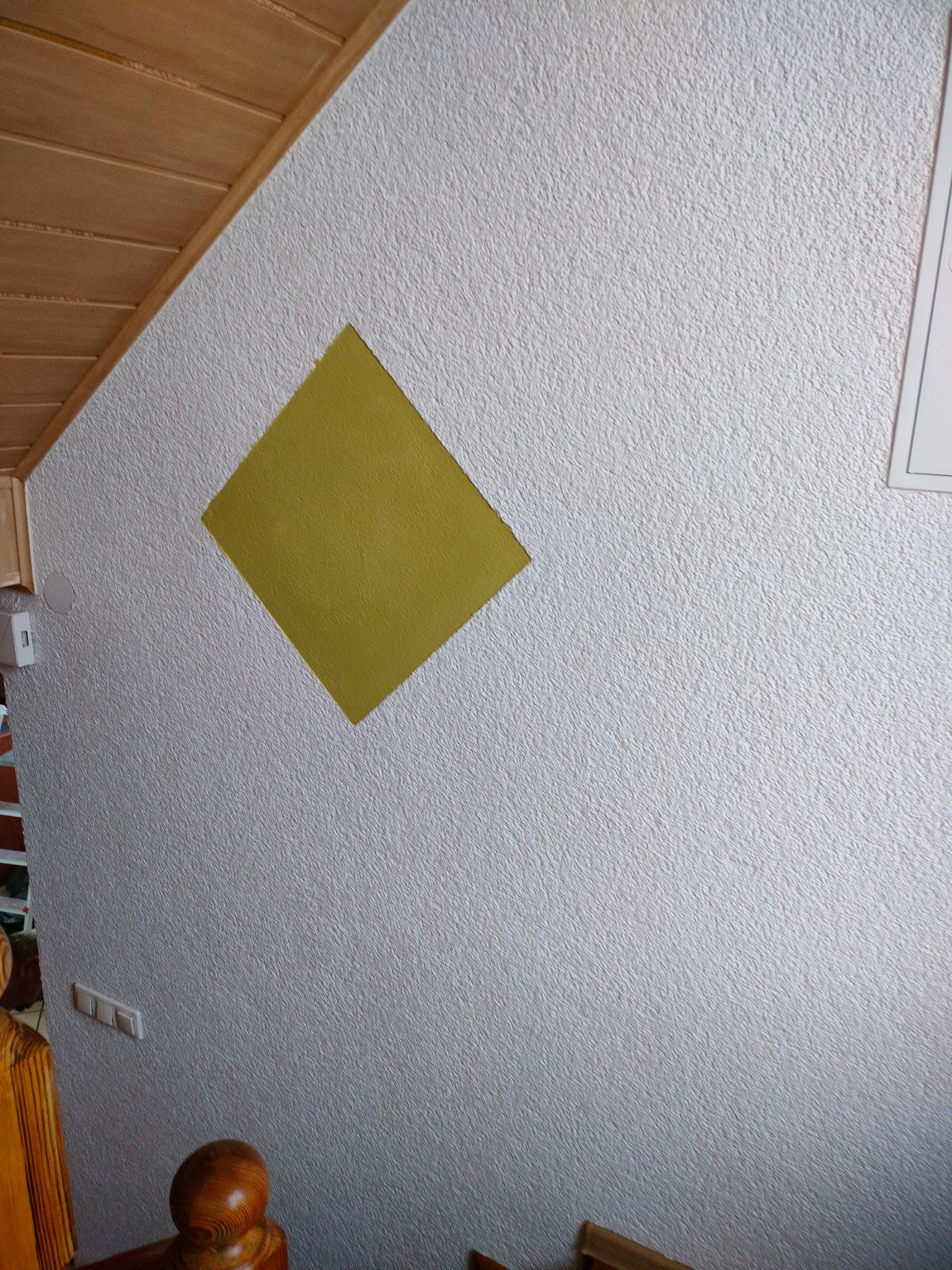 Fragment ściany z fakturą i pomalowanym żółtym rombem, widoczne włączniki, fragment drewnianego sufitu i balustrady.