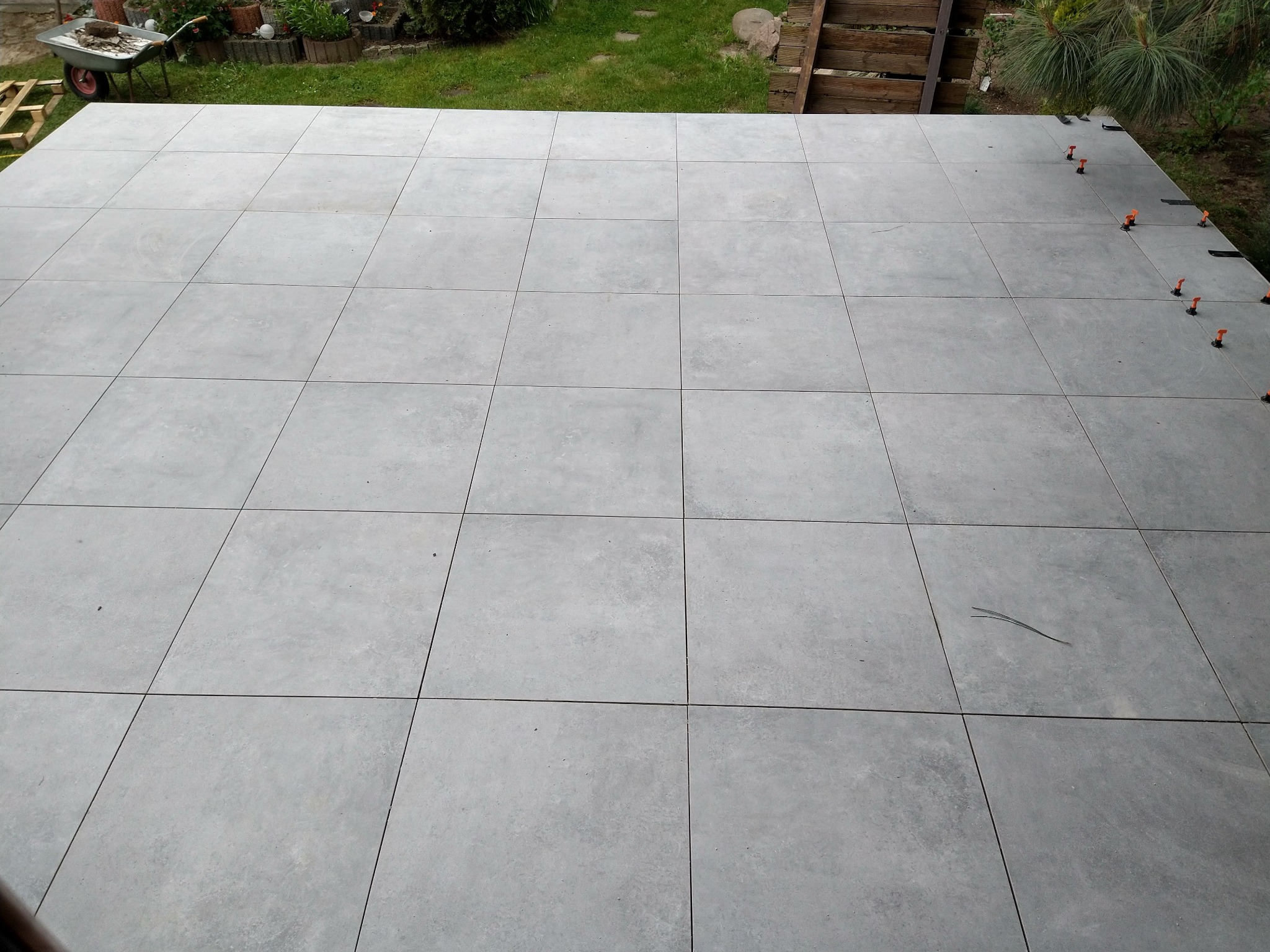 Układanie szarych, kwadratowych płytek na tarasie, widoczne pomarańczowe elementy poziomujące, w tle trawnik i drewniane ogrodzenie.