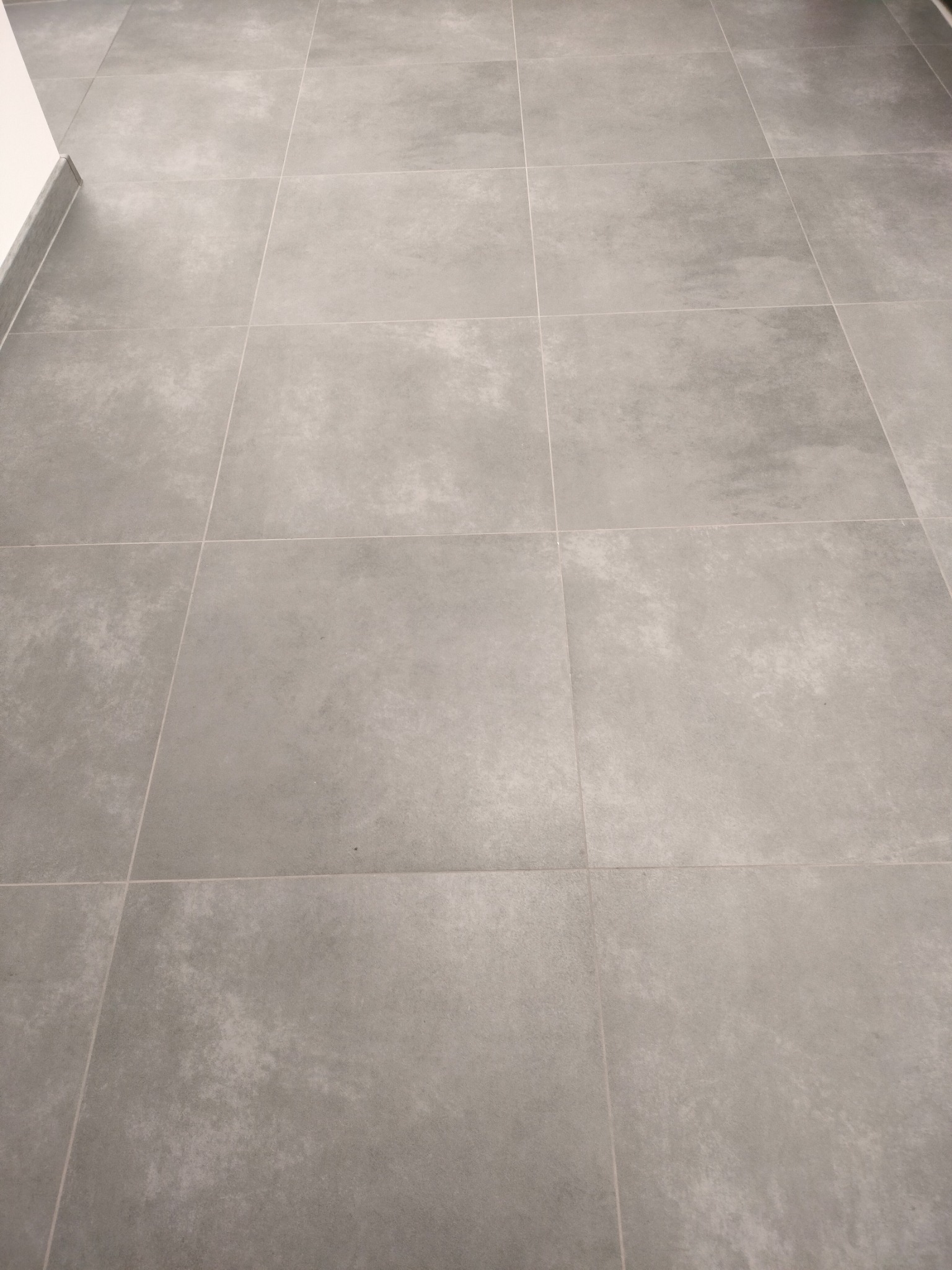 Szare, matowe płytki podłogowe ułożone w kwadraty z fugami, widoczne przy listwie przypodłogowej.